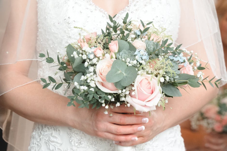 A bride holding a wedding bouquet composed of soft pink roses, white baby's breath, and eucalyptus leaves, arranged in a rounded, lush style. The flowers are fresh, with delicate petals and vibrant gr