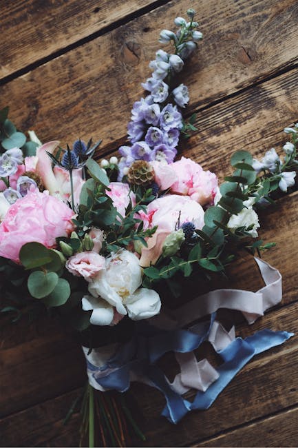 A floral bouquet featuring large, soft pink peonies, white garden roses, and sprigs of blue veronica flowers arranged with lush green eucalyptus leaves. The bouquet is wrapped with elegant satin ribbo